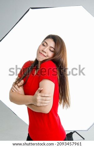 Portrait studio shot of Asian young happy cheerful female model in red casual shirt sitting on chair smiling look at camera posing crossed arms in front softbox lighting in photographing shooting set.