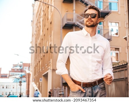 Portrait of handsome smiling stylish hipster lambersexual model.Modern man dressed in white shirt. Fashion male posing on the street background in sunglasses. Outdoors at sunset 
