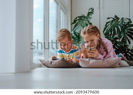 Children lie on the floor of the window and watch cartoons on their phones. Brother and sister play games on the phone.