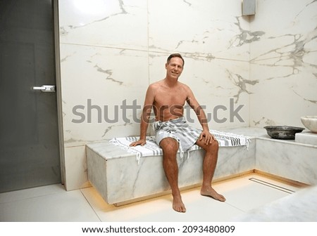 Handsome charming mature 50 years old European man wrapped in a towel sitting on a marble surface at Turkish hammam, getting relaxing male beauty treatment at luxurious spa wellness centre