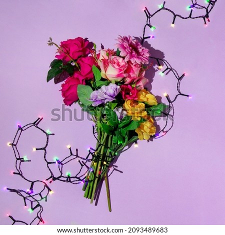 Creative layout with colorful bouquet of flowers and colorful christmas lights string on pastel purple background. 80s or 90s retro fashion aesthetic party concept. Minimal New Year celebration idea.