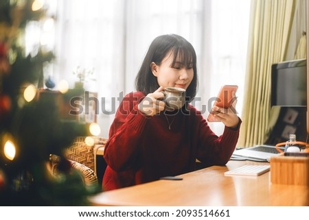 Young adult asian woman sipping a coffee and trade stock on mobile phone on morning. World trend crypto currency and nfts concept. Background at home office with computer.
