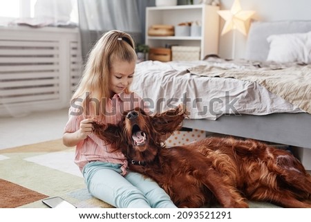 Portrait of cute blonde girl playing with Irish Setter dog on floor in cozy home interior, copy space