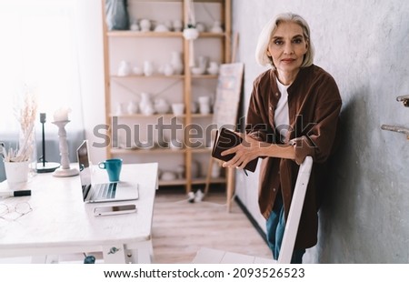 Serious senior caucasian female entrepreneur with notebook standing near working table and looking at camera. Small business and entrepreneurship. Modern successful woman. Interior of home art studio