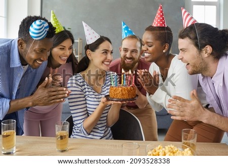 Group of cheerful young multiracial people in conical party hats applaud while the birthday girl makes a wish. Happy woman holding a birthday cake while celebrating at home with her best friends.
