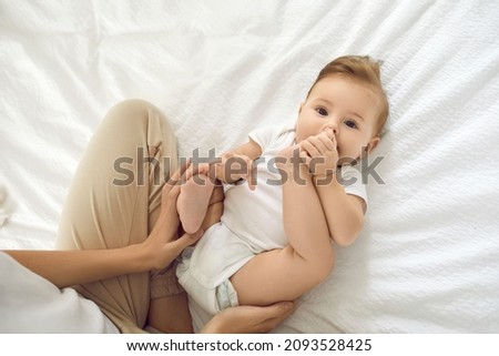 Portrait of cute charming six month old girl playing with her legs lying on her back on white cotton bed in bedroom. Baby in white bodysuit lying next to unrecognizable mother and looking at camera.