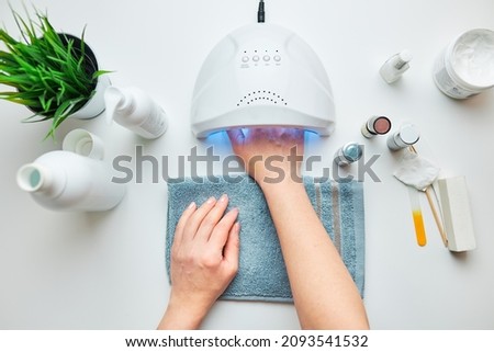Woman applying gel hybrid polish using UV lamp. Beauty wellness spa treatment concept. Cosmetic products, UV lamp, green leaves on white table. Spa, manicure, skin care concept. Flat lay, overhead Royalty-Free Stock Photo #2093541532