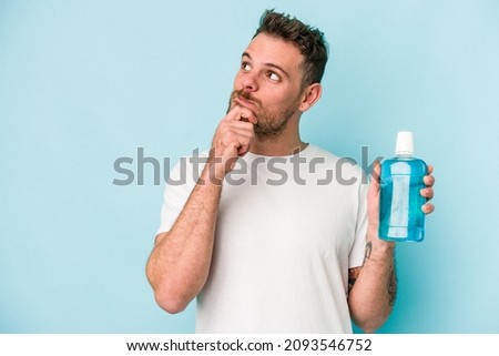 Young caucasian man holding mouthwash isolated on blue background looking sideways with doubtful and skeptical expression. Royalty-Free Stock Photo #2093546752
