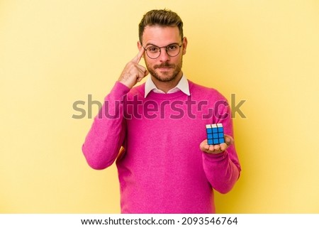 Young caucasian man holding Rubik’s cube isolated on yellow background pointing temple with finger, thinking, focused on a task. Royalty-Free Stock Photo #2093546764