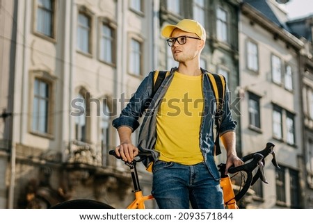 Portrait of a happy delivery man who is sitting on a bicycle. He is wearing yellow clothing. He is carrying a yellow backpack on his shoulders. Center of the old city.