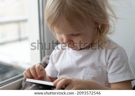 Little girl with blonde hair is sitting on the window and using her phone close up.