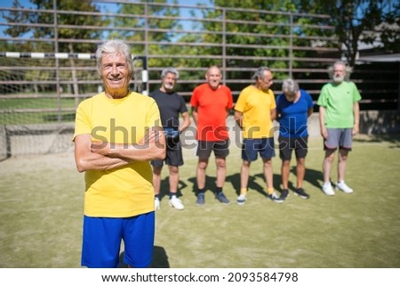 Portrait of happy senior man on football field. Captain with gray hair in sport clothes standing, looking at camera, teammates in background. Football, sport, leisure concept