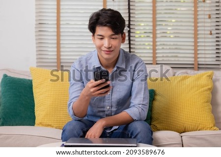 Sitting on the sofa in the house, an Asian man works on his smartphone.Man works while using social media applications. The concept of working from home.