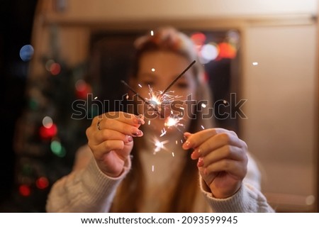 sparklers bengal fire stick in female hands holding woman in knitted wool white cozy sweater on the background of a camping trailer house outdoor on new year and christmas eve night. sparkling sparks