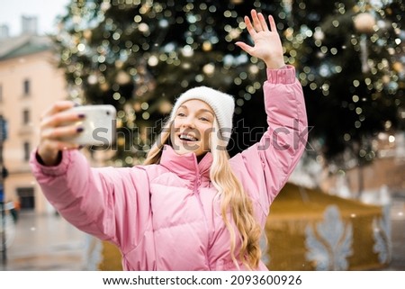 A girl in the winter makes a photo on the background of a Christmas tree on the phone. Roaming communication during the holidays, the concept of communication with loved ones. New Year, smile
