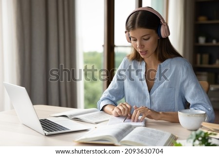 Focused engaged student girl in big headphones studying foreign language, listening audio lesson at laptop, reading notes out loud, doing exercises from open book, school, college homework task Royalty-Free Stock Photo #2093630704