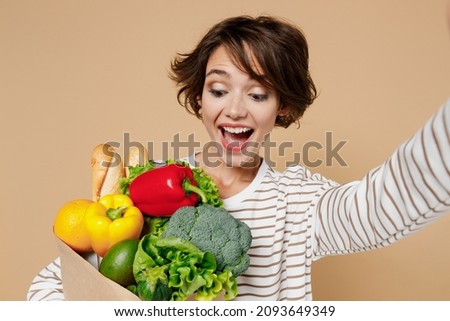 Close up young amazed happy fun vegetarian woman 20s in casual clothes hold paper bag with vegetables doing selfie shot pov on mobile phone isolated on plain pastel beige background Shopping concept