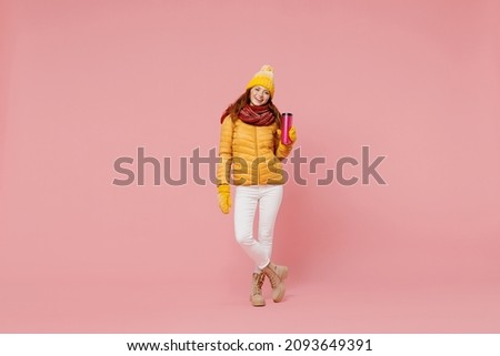 Full size body length jubilant fascinating exultant young woman 20s years old wears yellow jacket hat mittens hold thermal cup coffee tea isolated on plain pastel light pink background studio portrait
