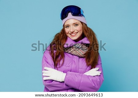 Skier happy satisfied smiling woman wear warm purple padded windbreaker jacket ski goggles mask spend extreme weekend in mountains hold hands crossed folded isolated on plain blue background studio.