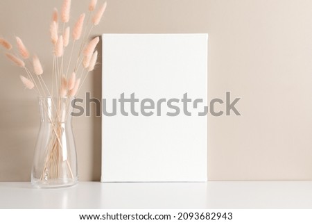 Blank white canvas mockup and bouquet of fluffy plants with pom-poms in glass vase on beige background. 