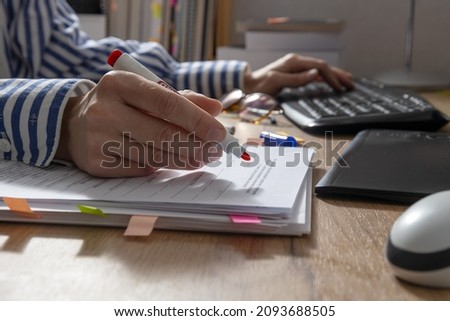 Closeup of woman hand who holding red marker and cheking text for grammar and using computer.Office table and documents,keyboard,mouse,tablet,pile of books on it