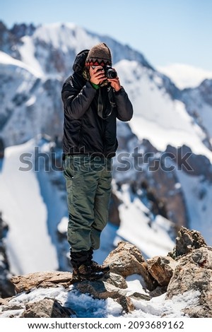 Young man trekker photographer taking pictures with camera on top of Pik Uchitel peak . Ala Archa Alpine National Park Landscape near Bishkek, Tian Shan Mountain Range, Kyrgyzstan, Central Asia.