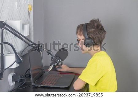 A teenage boy in a yellow T-shirt is blogging at home in front of a laptop with a microphone and headphones.