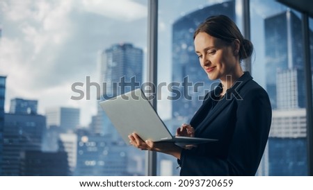 Successful Businesswoman in Stylish Dress Working on Laptop, Standing Next to Window in Big City. Confident Female CEO Analyze Financial Projects. Manager at Work Planning Marketing Campaign. Royalty-Free Stock Photo #2093720659