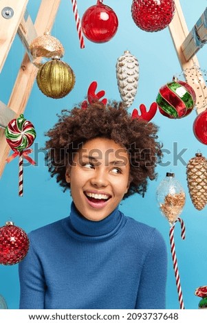 Happy woman with curly hair looks away gladfully smiles toothily going to celebrate New Year decorates room before festive event poses against toys isolated over blue background. Holiday decoration