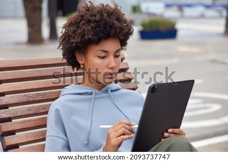 Photo of serious woman concentrated at tablet screen draws pictures with stylus poses on wooden bench dressed in hoodie spends leisure time outdoors against blurred background works freelance