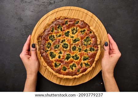 Women's hands hold quiche pie with broccoli, tuna and cheese on round wooden cutting board on black graphite background. Top view  Royalty-Free Stock Photo #2093779237