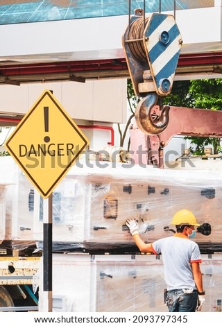 back of asian male worker working under a large overhead hook with yellow danger warning sign