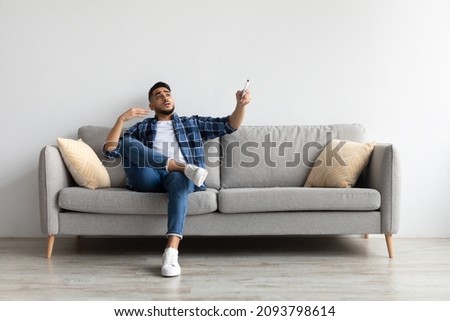 Split System. Portrait Of Middle Eastern man suffering from summer heat, sweaty guy holding remote control switching on ac air conditioner, sitting on couch in living room cooling himself waving hand Royalty-Free Stock Photo #2093798614