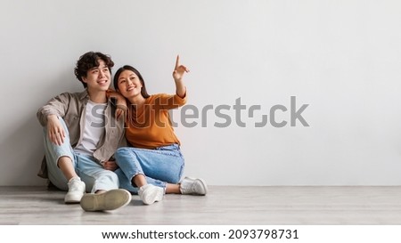 Carefree millennial Asian man and woman sitting on floor near white studio wall and pointing at copy space, banner design. Young couple offering place for cool advertisement, panorama Royalty-Free Stock Photo #2093798731