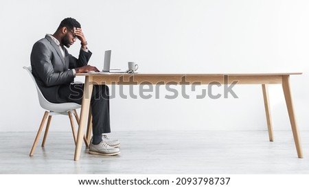 Side view of young black office employee feeling tired of working overtime, sitting at desk with laptop against white studio wall, copy space. Exhausted businessman having problem with online job Royalty-Free Stock Photo #2093798737