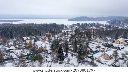 Winter rural landscape. Aerial view of a snow-covered village near the lake. Top view of houses and trees. Cold snowy winter weather. Snowfall. Snow on the roofs. Toksovo, Leningrad region, Russia. Royalty-Free Stock Photo #2093801797