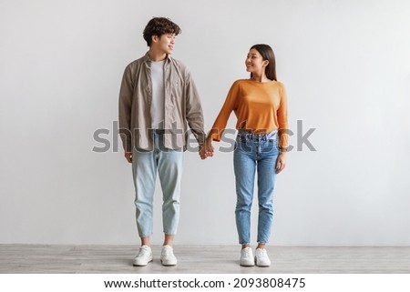 Full length portrait of millennial Asian couple smiling and looking at each other, holding hands, standing against white studio wall. Affectionate young man and woman being in love Royalty-Free Stock Photo #2093808475