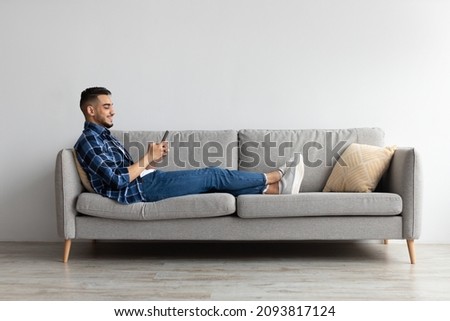 Cool Gadget And Application. Portrait of young smiling Arab man holding mobile phone, typing sms message, sitting on the sofa in living room. Guy browsing internet, surfing web, using app, free space Royalty-Free Stock Photo #2093817124