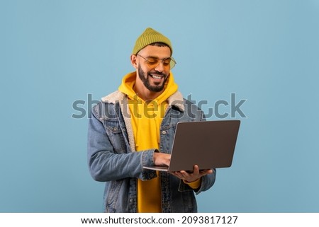 Happy young arab freelancer guy with laptop working online and browsing internet standing over blue studio background. Technology, great website concept Royalty-Free Stock Photo #2093817127
