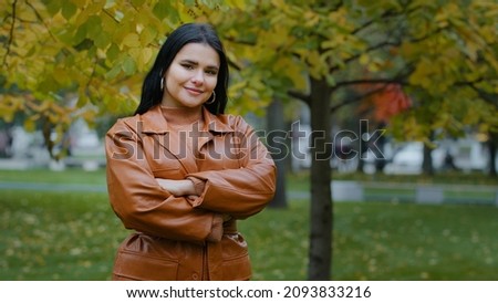 Beautiful young elegant woman looking at camera focuses gaze crossed arms over chest smiles shyly posing attractive pretty Hispanic girl stand in city autumn park outdoors portrait happy female