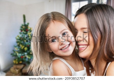 Beautiful mother and daughter have fun, hug and kiss at home near the Christmas tree in the interior.