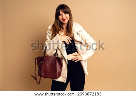 Stylish young brunette woman posing at fur coat, beige studio background, winter time. trendy accessorizes. Royalty-Free Stock Photo #2093872183