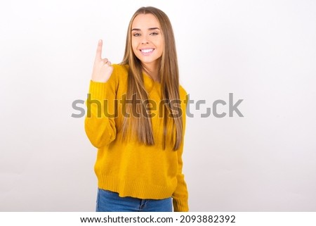 young caucasian woman wearing yellow sweater isolated over white background smiling and looking friendly, showing number one or first with hand forward, counting down