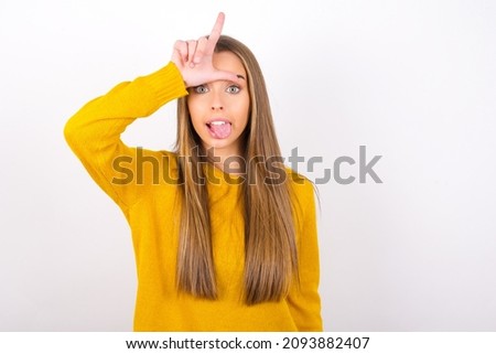young caucasian woman wearing yellow sweater isolated over white background gestures with finger on forehead makes loser gesture makes fun of people shows tongue