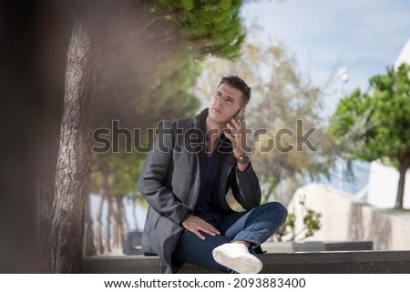 Classy freelance architect sitting alone on a bench outdoor and talking on the phone during a beautiful summer day. Classy small entrepreneur using the smart phone along the riverside in Lisbon Royalty-Free Stock Photo #2093883400