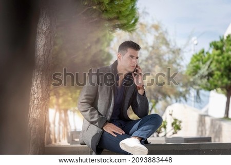 Classy freelance architect sitting alone on a bench outdoor and talking on the phone during a beautiful summer day. Classy small entrepreneur using the smart phone along the riverside in Lisbon Royalty-Free Stock Photo #2093883448