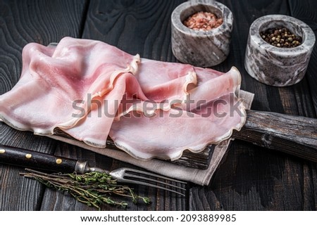 Sliced prosciutto ham on wooden board with herbs. Black wooden background. Top view