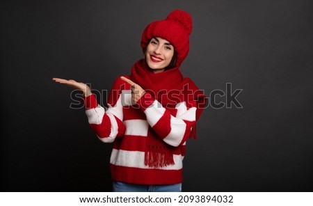 Close up portrait of a beautiful smiling girl in winter hat and scarf while she pointing on her palm isolated on gray background