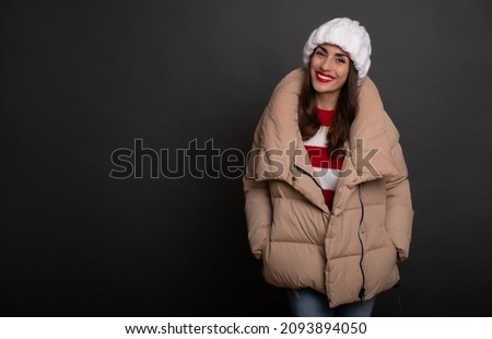 Close up portrait of a beautiful smiling woman in a winter hat and warm stylish puffer while she posing and looking on camera isolated on gray background