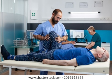Physiotherapist helping old man with leg injury to recover at kinetotherapy clinic. Chiropractor giving support to retired patient with knee pain and muscle pressure for recovery. Royalty-Free Stock Photo #2093918635